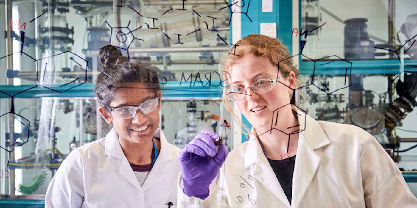 Two researchers writing chemical formulas on a transparent screen