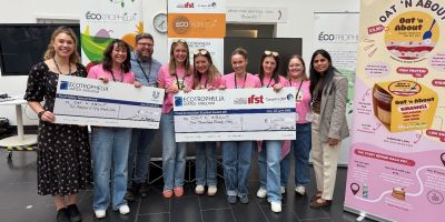 The winning team with their oversized prize money cheques, standing in front of some banners advertising Ecotrophelia. The team, all wearing pink t-shirts, are joined by Leeds staff Amie Lister, Gleb Yakubov and Afreen Rahman. Everyone is smiling and look