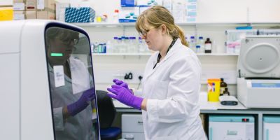 A food science technician in front of an incubator in the lab. They're wearing a white lab coat and purple gloves.