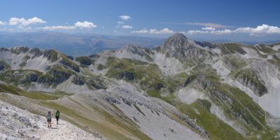 A landscape view of the Gran Sasso d'Italia mountain range. To the left, two people are hiking on a stony path.
