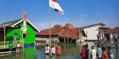 A flooded village with small buildings coming out of the water. An Indonesian flag is in the centre, with people standing in the floodwater around it.