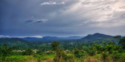 A lush green landscape with mountains far in the distance. The sky is covered in clouds which get darker as they get further away.