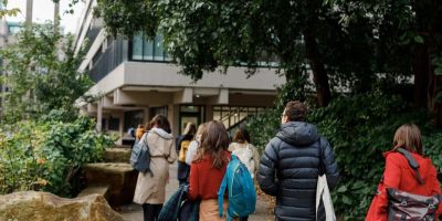 Students walking along a paved area towards a brutalist style building. Bushes and trees on either side.