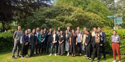 A large group of international researchers standing on the grass at the University of Leeds campus with trees and bushes behind them.
