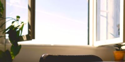 An open window with a blue sky, sun coming in to the room with a plant and a desk.