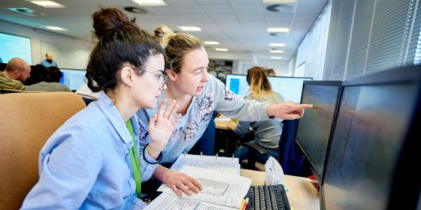 A girl sits at a desk with computer monitors, keyboard and papers while another woman points something out on screen