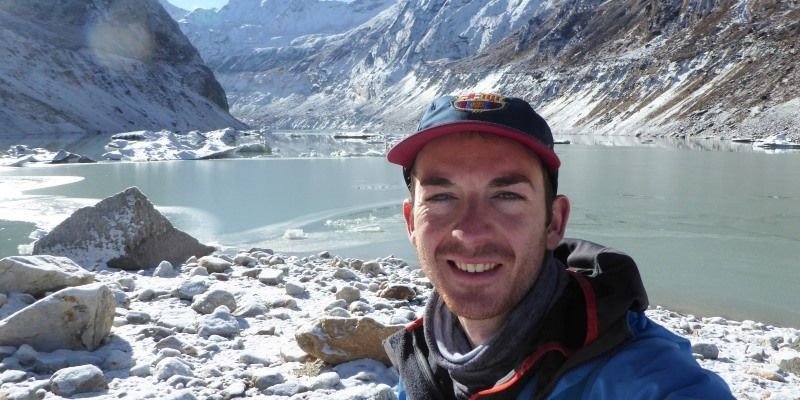 Dr C Scott Watson wearing a coat and cap, in front of a glacial lake.